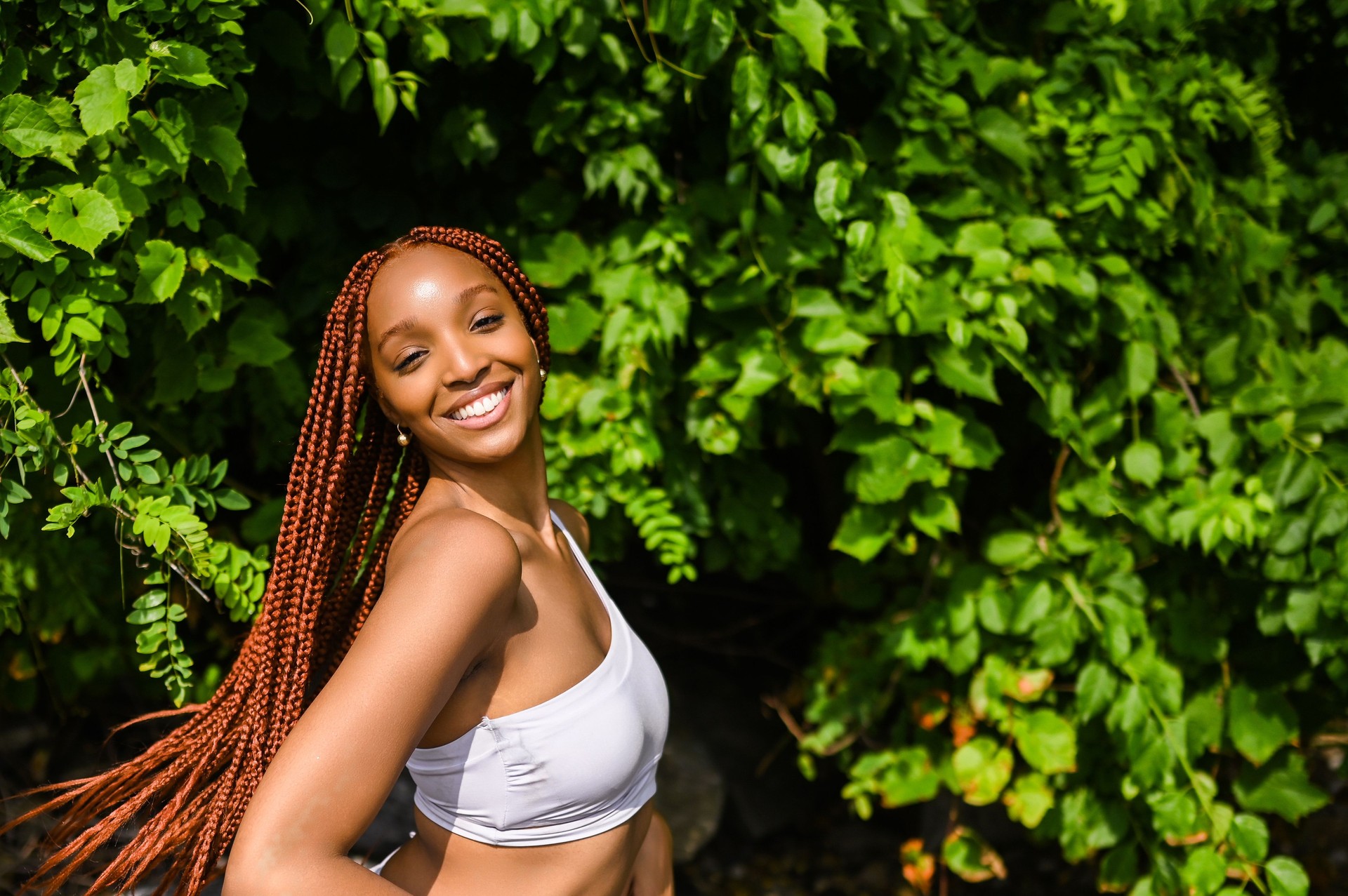 Outdoor portrait of natural Beautiful young African American woman long red braids hair style and perfect white teeth smile, posing in swimsuit at sunny summer day with green foliage beach background.
